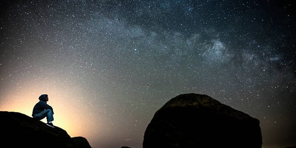 Man sitting on the rocks in solitude under the cover of dark, clear sky and looking at Milky Way galaxy while thinking about his existence.